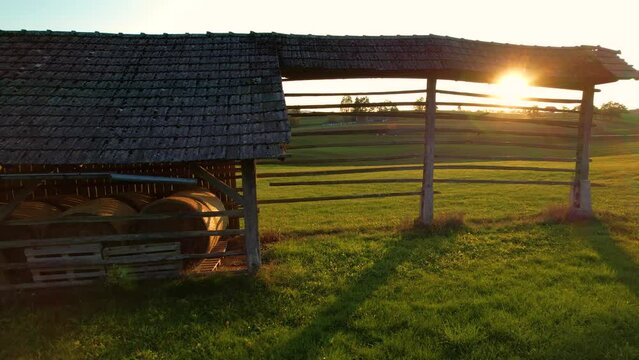 LOW ANGLE: Sunbeams shining through traditional hayrack with stored hay bales. Old wooden farm building for hay drying and storage standing in the middle of beautiful green pasture in golden light.