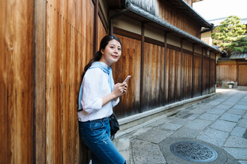 cheerful Taiwanese lady leaning against historic wooden wall with smartphone in hand is smiling while looking into distance at Ishibe Alley in gion, kyoto japan