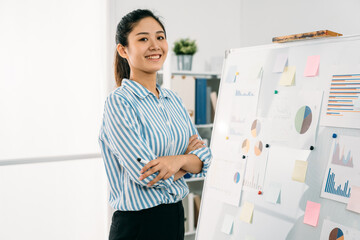 confident asian businesswoman standing by whiteboard with graphs and charts in striped shirt is...