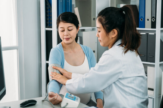 portrait female medical doctor is wrapping the cuff around the expectant patient’s arm to monitor her blood pressure during pregnancy checkup in the hospital.