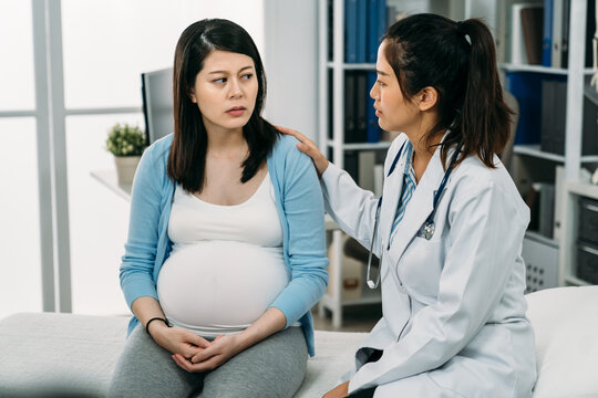 Asian Pregnant Woman Suffering Depression Is Looking At Her Psychologist Doctor Who Is Patting On Her Shoulder Showing Support During Consultation In The Clinic