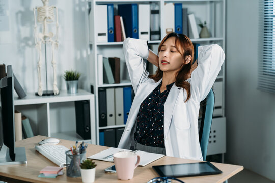 portrait of asian female physician is putting her hands behind head and closing her eyes to relax while taking a break from work at desk on the office chair.