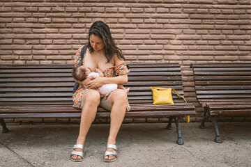 young mom breastfeeding newborn baby daughter in public outdoors in the street on a bench - obrazy, fototapety, plakaty