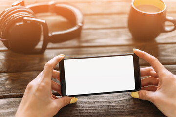 Female hands holding phone with place for text on a screen at wooden table near a cup of coffee and headphones. Point of view in first person.