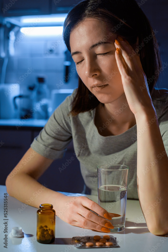 Wall mural A young housewife woman is sitting at the kitchen table at night with a headache. There are pills and a glass of water nearby.