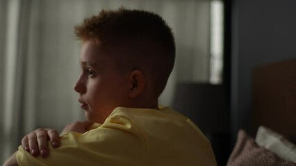 Close-up face of sad unhappy boy hugging knees and crying sitting on bed in dark bedroom on background of window. Lonely redhead child putting head down on knees, thinking over childish problems.