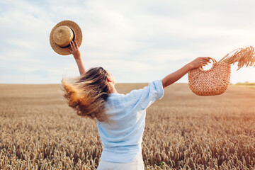 Back view of young happy woman jumping enjoying view in summer field holding straw handbag with...