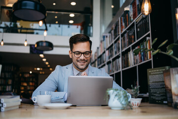 Young handsome man sitting in modern bookstore, reading and using his laptop or notebook computer.