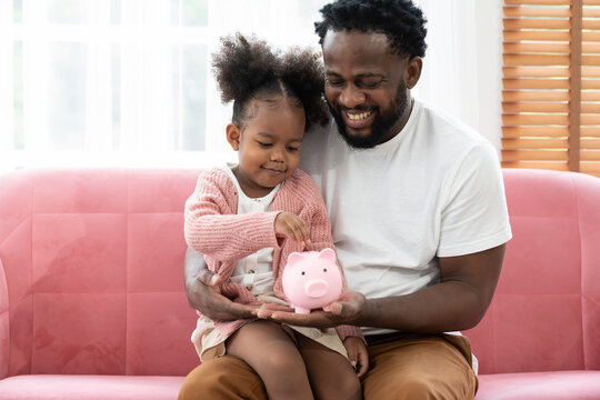 Father And Daughter Inserting A Coin In A Piggy Bank. Happy African American Small Girl Kid Putting Money Into The Piggybank