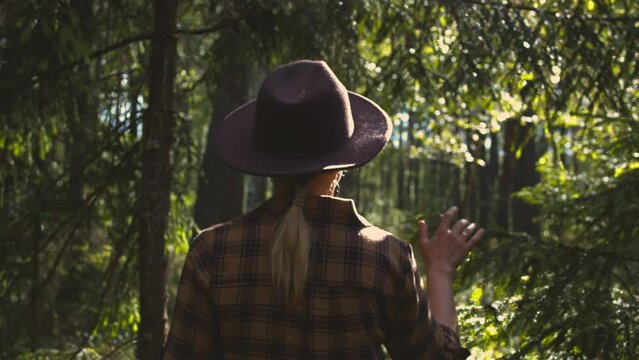 A young blonde woman in a plaid shirt and a brown hat walks slowly through a sunny forest.advertising concept for travel,hiking,hiking and travel clothing. Healthy lifestyle.