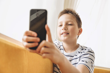 Teenage boy sitting on couch using smartphone texting and having video chat on internet online at home. Child holding mobile phone and looking at screen. Social media. Young blogger influencer