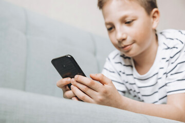 Teenage boy lying on couch using smartphone and playing video games on internet online at cozy home. Child holding mobile phone and looking at screen. Social media