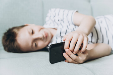 Teenage boy lying on couch using smartphone and playing video games on internet online at cozy home. Child holding mobile phone and looking at screen. Social media