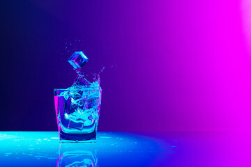 Ice cube falls into a transparent glass of water standing on mirror surface over gradient blue-pink...