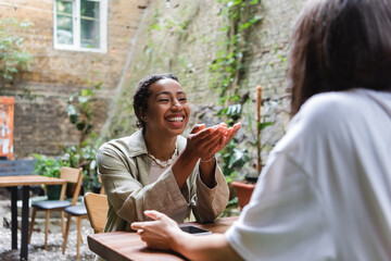 Cheerful african american woman talking to friend near smartphone in outdoor cafe
