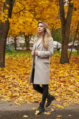 young beautiful woman with long blond hair in a light coat in the park in autumn against the background of trees with yellow leaves