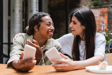 Cheerful african american woman holding blurred cellphone near friend in outdoor cafe