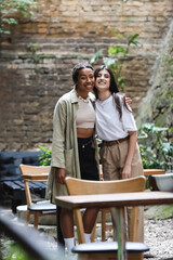 Fototapeta premium Smiling interracial girlfriends hugging and looking at camera on terrace of cafe