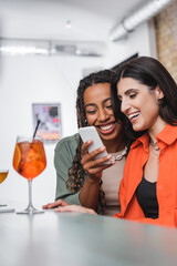 Smiling african american woman using smartphone near friend and blurred cocktails in cafe