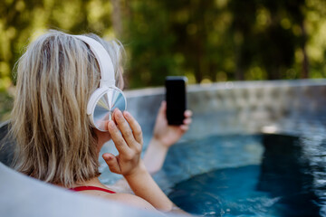 Reaxed woman with headphones listening to music in outdoor bathtub, rear view.