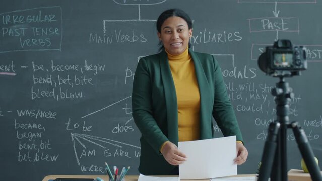 African American woman teaching English in classroom and recording lesson with video camera. Chalkboard with grammar rules visible in backgound.