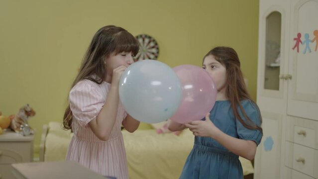 Little girls are blowing air into the balloon. Happy birthday party, celebration. Children are having fun by inflating balloons in their rooms. Cute kids inflate the balloon for their birthday.