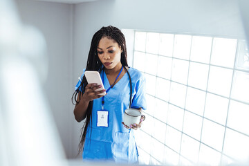 African female surgeon wearing scrubs reading text message on her mobile phone while standing in...