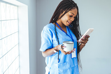 African female surgeon wearing scrubs reading text message on her mobile phone while standing in...
