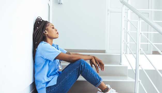 Female Nurse Sitting On The Floor And Looking Distraught. Exhausted Sad Black Doctor Feels Burnout Stress. Stressed Female Medical Taking Break From Work. Being An Intern Is Tough Work