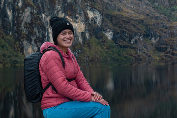 Mujer alegre con mochila sentada en una roca contra un lago rodeado de montañas