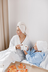 Mom and her little daughter in bathrobes and towels on their heads drink tea and eat pizza in bed. 