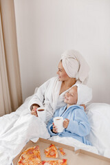Mom and her little daughter in bathrobes and towels on their heads drink tea and eat pizza in bed. 