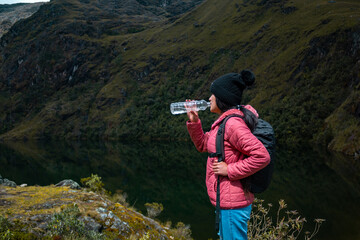 Caminante en una chaqueta rosa bebiendo agua junto a un lago tranquilo rodeado de montañas...