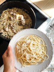 Homemade pasta with cheese and meat in pepper sauce in a frying pan. The hand holds a white plate with a dish