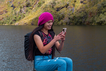 Excursionista atractivo sentado y sosteniendo un teléfono junto a un lago tranquilo en las...