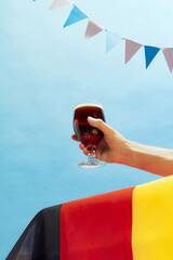 Hand with glass of cold dark beer over light blue sky and white-blue flags background. Oktoberfest,...