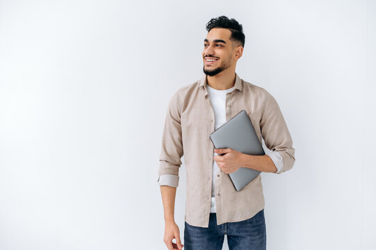 Positive friendly modern arabian or indian young man with beard, wearing casual shirt, standing on isolated white background, holding laptop, looking to the side, smiling, dreaming, thinking
