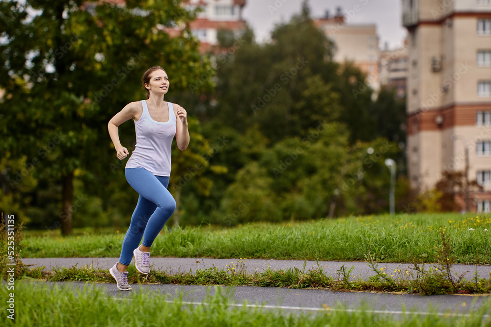 Wall mural an athlete woman warms up by jogging in summer city park near residential area with multi apartment 