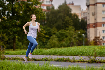 An athlete woman warms up by jogging in summer city park near residential area with multi apartment...