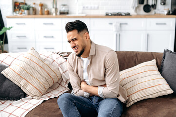 Frustrated unhealthy indian or arabian young man, sitting on the sofa in the living room, holding his stomach, suffering from spasm, stomach pain, poisoning, needs treatment, sad facial expression