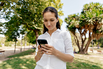 Adorable lobby woman with long dar hair in whit shirt walking in summer green park with smartphone....