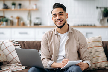 Handsome friendly indian or arabian man taking notes while working on project or learning online, listening webinar uses laptop, sitting on a sofa in a living room, prepares a report, looks at camera
