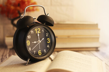 Black alarm clock, in front of a pile of books, on a wooden table and a white textured wall...