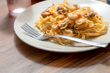 A white plate on a wooden table with pasta with mussels and shrimp, a fork and a glass of water.