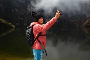 Chica con una chaqueta rosa con una mochila parada junto al lago en las montañas y tomando selfies - obrazy, fototapety, plakaty