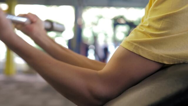 The guy with thin skinny body working hard in street sports ground. The guy trains his biceps with an empty barbell in a fixed position. Workout, sports lifestyle, thin athlete.