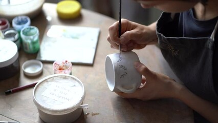Hands of woman hold white ceramic mug painting creative patterns with brush in pottery studio. Female artisan in black apron sits at round table near equipment enjoying handmade craft close view