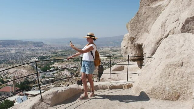 Tourist woman in straw hat takes a photo on the phone in valley in Cappadocia in Turkey. Girl making video  of beautiful landscape with mobile camera, dreamy travel concept.