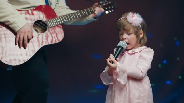 Little girl on stage in vintage dress, she sings into microphone and dances, her father plays acoustic guitar. Color music is shining. Performance on stage. Early development of children