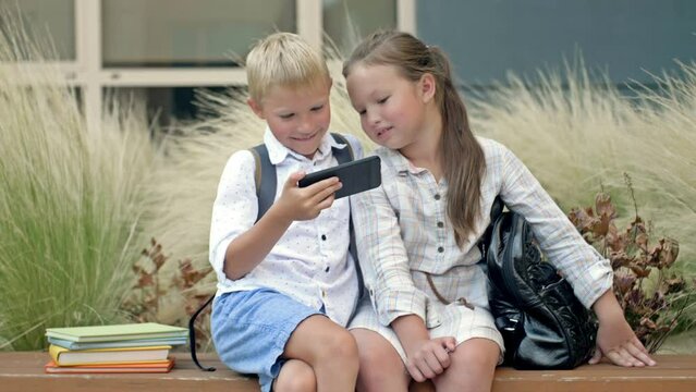 Two friends, a boy and a girl aged 8-9, are sitting on a bench in the schoolyard. Children look at the smartphone screen with interest.